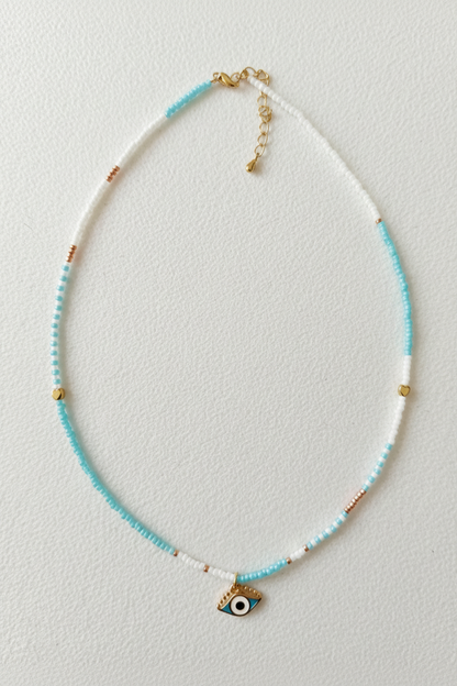 A delicate beaded necklace with white and light blue beads, accented with small gold beads and a small evil eye charm at the center, displayed on a textured off-white background.