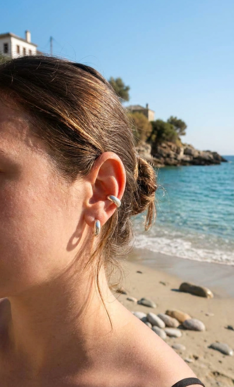 A close-up side profile of a woman with a small silver hoop earring in her ear, photographed outdoors on a sandy beach with blue sea and sky in the background.